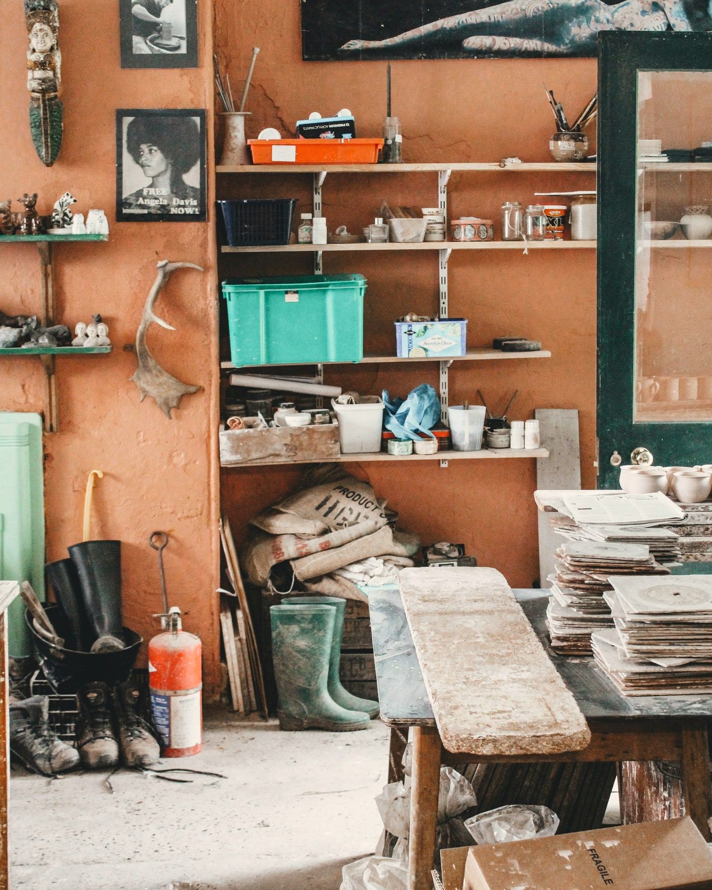 A studio wall of shelves and hanging tools above a worn workbench, stacked with jars, pots and materials.